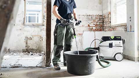 Une artisane se tient sur un chantier et utilise un malaxeur Festool en combinaison avec un aspirateur.